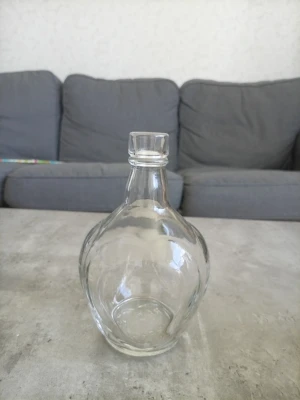 Genomskinlig glasflaska med kork - Säljer en stilren, rundad glasflaska i genomskinligt glas med tillhörande glaspropp. Perfekt för att förvara drycker, oljor eller som dekoration. Flaskan har en bred botten och smal hals vilket ger den en klassisk look.