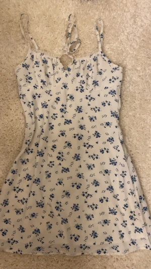 Vit blommig kortklänning med snörning - Söt vit kortklänning med blå blommigt mönster och smala axelband. Klänningen har snörning framtill vid bysten och en lätt utsvängd passform. Perfekt för varma dagar och ger en fräsch sommarkänsla.