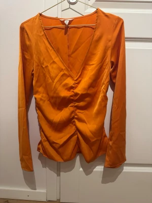 Orange v-ringad topp från H&M - Säljer en snygg orange långärmad topp från H&M med v-ringning och rynkade detaljer framtill. Toppen har en figurnära passform och är tillverkad i ett glansigt syntetmaterial som ger en cool look.