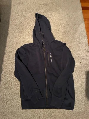 Marinblå hoodie från Sail Racing - Snygg marinblå hoodie från Sail Racing med dragkedja framtill och logga på bröstet. Hoodien har huva med dragsko, ribbade muddar och mjukt fleecefoder på insidan. Perfekt för dig som gillar sportig och avslappnad stil. Riktigt schysst tröja