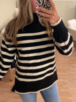 Svartvit randig stickad tröja - Snygg stickad tröja med svarta och vita horisontella ränder och hög krage. Tröjan har långa ärmar och ribbstickade muddar. Perfekt för kyliga dagar och enkel att matcha med jeans eller kjol.