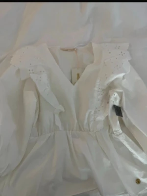 Vit blus med volang och broderi - Söt vit blus med v-ringning, volangdetaljer och broderade hålmönster på axlarna. Blusen har lång ärm och markerad midja för en feminin siluett. Perfekt för dig som gillar romantisk och trendig stil.