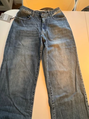 Blå wide jeans med blixtdetalj på bakfickan  - Säljer ett par klassiska blå jeans med vid passform och fem fickor. Jeansen har en cool gul blixtdetalj på ena bakfickan och slitna kanter på fickorna. Perfekta för en avslappnad och trendig look. Nya med lappar på från Design by Si. Storlek XS, midjemått 31,5cm, ben innelängd 82cm 