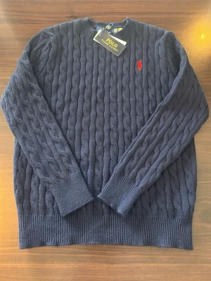 Mörkblå kabelstickad tröja Polo Ralph Lauren - Kabelstickad mörkblå tröja från Polo Ralph Lauren med rund halsringning och röd broderad logga på bröstet. Tröjan har ribbade muddar vid ärmslut och nederkant, och är tillverkad i mjukt material som ger en klassisk och stilren look.