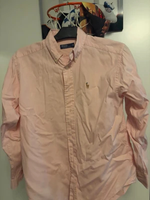 Ljusrosa skjorta från Polo Ralph Lauren - Säljer en klassisk ljusrosa skjorta från Polo Ralph Lauren med broderad logga på bröstet. Skjortan har lång ärm, knappar framtill och en tidlös passform. Perfekt för dig som gillar stilrena plagg med ikonisk detalj.