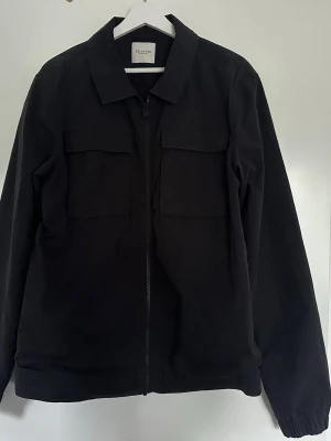 Svart overshirt från Selected Homme - Stilren svart overshirt från Selected Homme med dragkedja framtill och två stora bröstfickor med lock. Klassisk krage och raka ärmar. Perfekt lager-på-lager-plagg för en clean look. Materialet ser ut att vara bomull eller bomullsblandning.