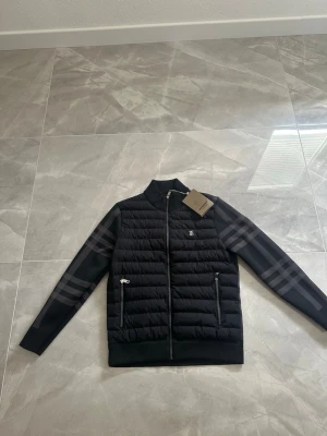 Svart pufferjacka från Burberry - Snygg svart pufferjacka från Burberry med klassiskt rutmönster på ärmarna och silverfärgad dragkedja. Jackan har två fickor med dragkedja och Burberrys ikoniska TB-logga på bröstet. Perfekt för kalla vinterdagar och riktigt stilren look.
