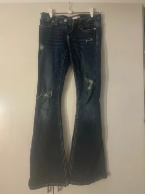 Blå bootcut jeans - Säljer ett par blå bootcut jeans med slitna detaljer och dekorativa sömmar på bakfickorna. Jeansen har låg midja och klassisk femficksmodell. Materialet är denim med stretch för extra komfort. Perfekta för dig som gillar en avslappnad och trendig look. Du ser på några bilder var de finns slitage 