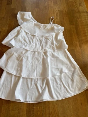 Vit oneshoulder volangklänning - Superfin vit kortklänning med volanger och oneshoulder-design. Klänningen har tunna axelband på ena sidan och bred volang över axeln. Perfekt för sommarens varma dagar och har en luftig passform.