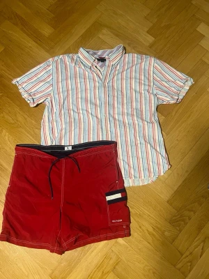 Tommy Hilfiger Randig Skjorta & Badshorts - Säljer det ultimata sommarsetet från Tommy Hilfiger, Perfekt för stranden, poolhäng eller en fräsch sommaroutfit.  Skjortan XXL Badbyxorna L   Obs! Shortsen har en väldigt liten, knappt synlig fläck på ena låret.