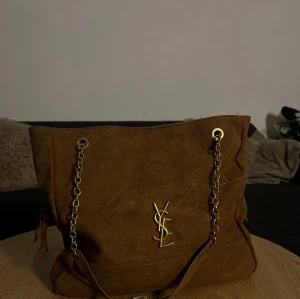 Ysl Brun  handväska med kedjerem - Snygg brun handväska i  från Yves Saint Laurent med guldfärgade kedjeremmar och YSL-logga framtill. Väskan har en mjuk, rymlig form och detaljer i guld som ger en lyxig känsla. Perfekt för dig som vill ha en trendig accessoar.