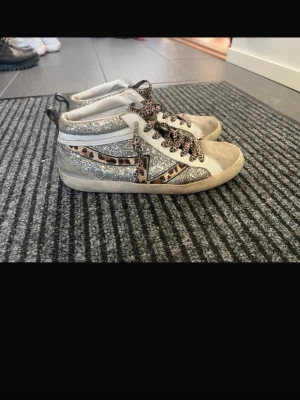 Glittriga sneakers med leoparddetaljer - Unika sneakers i mid-modell med silverglittriga paneler, beige mocka framtill och leopardmönstrade snören samt detaljer på sidan. Skorna har vit bas och en beige sula som ger en cool kontrast. Perfekta för dig som vill sticka ut med din stil.