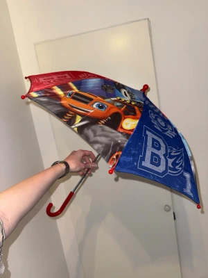 Färgglad barnparaply med bilar - Säljer ett färgglatt paraply för barn med motiv av tecknade bilar i rött, blått och svart. Paraplyet har ett böjt rött handtag..