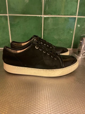 Lanvin skor  - Snygga svarta sneakers med vit sula och lackad tå. Skorna har svarta skosnören, metallöljetter och en stilren design. Ovandelen är i mockaliknande material medan tån är i blankt lack. Perfekta för en trendig och avslappnad look.