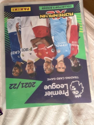 Premier League set - Säljer ett Premier League Adrenalyn XL samlaralbum från säsongen 2021/22 med tillhörande samlarkort. Albumet har färgglad framsida med spelare från olika lag och medföljer en stor hög med fotbollskort i olika färger och motiv.