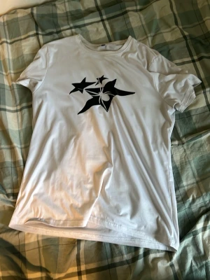 Vit t-shirt med svart stjärntryck - Snygg vit t-shirt med ett stort svart tryck av stjärnor på bröstet. Klassisk passform och rund halsringning. Perfekt till jeans eller shorts för en clean och stilren look.