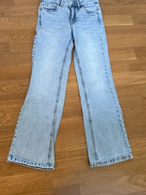 Ljusblå  jeans med knappfickor i bak  - Säljer ett par ljusblå jeans med knappfickor i bak. Jeansen har klassiska fem fickor, bälteshällor och de har resårband i midjan (se bild 3). Materialet är denim i bomull och benen har en lätt utsvängd siluett. Perfekta för en avslappnad och trendig look.