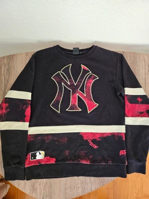 Svart långärmad New York topp - Cool svart långärmad topp med stort New York-tryck fram och texten 'NEW YORK' bak. Röd och vit detalj på ärmar och nederkant ger en sportig vibe. Perfekt för dig som gillar streetwear och vill sticka ut. Denär få använd och är i utmärkt skick. Storleken är Medium men passar också till Small i vissa fall.
