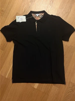 Svart pikétröja från Burberry - Klassisk svart pikétröja från Burberry med den ikoniska rutiga detaljen vid knäppningen. Tröjan har korta ärmar, krage och knappar framtill. Perfekt för en stilren och tidlös look.