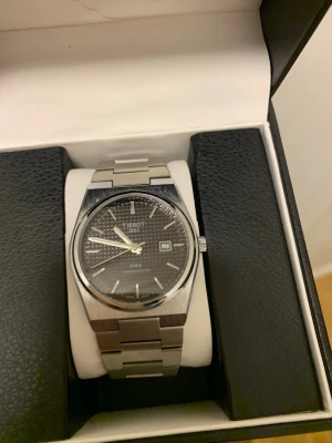 Tissot PRX klocka i silver - Snygg Tissot PRX klocka med silverfärgad länkarmband och svart urtavla. Klockan har datumvisning och stilrena indexmarkeringar. Perfekt accessoar för dig som gillar klassisk och modern design i ett.