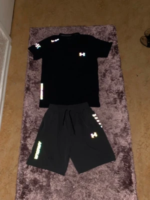 Under Armour Reflective-set - Säljer ett svart träningsset från Under Armour med t-shirt och shorts. Setet har reflekterande loggor och detaljer i regnbågsfärger. Materialet är lätt och snabbtorkande, perfekt för träning eller gym. Både tröjan och shortsen har en sportig passform och dragsko i midjan.