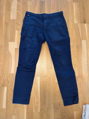 Mörkblå chinos med smal passform - Snygga mörkblå chinos med smal passform och klassiska sidfickor. Byxorna har bälteshällor och är tillverkade i ett mjukt bomullstyg som ger en skön känsla. Perfekta för en stilren och avslappnad look.Blåa chinos byxor storlek 32  Uppsydda för att passa mig 180cm Sommarbyxor Midja 45 cm Längd 100 cm