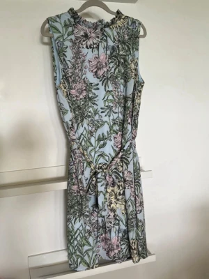 Blommig ärmlös kortklänning - Superfin ljusblå kortklänning med blommigt mönster i grönt, rosa och gult. Klänningen är ärmlös och har ett knytband i midjan för en snygg passform. Perfekt för varma dagar och somriga vibes! Storlek 44