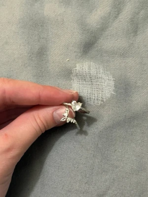 Silverfärgad ring med hjärtformad sten - Säljer en snygg äkta silver ring med en stor hjärtformad klar sten i mitten och mindre stenar längs bandet. Ringen har en unik design där två ringar sitter ihop och ger en extra glittrig look. Perfekt för dig som gillar statement-smycken.