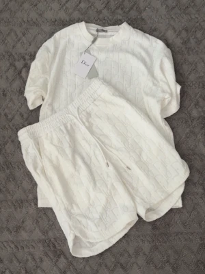Vitt set från Dior med mönster - Säljer ett lyxigt vitt set från Dior med diskret mönster i tyget. Setet består av en t-shirt med rund hals och ett par shorts med resår i midjan och dragsko. Materialet känns mjukt och bekvämt, perfekt för en avslappnad look.