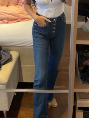 Blå high waist jeans med raka ben - Säljer ett par blå high waist jeans med raka ben och fyra synliga knappar framtill. Jeansen har klassiska fickor fram och bak samt bälteshällor. Materialet är denim i bomull och passformen är tight upptill och rak nedtill.
