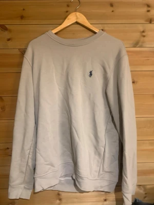 Ljusgrå sweatshirt från Polo Ralph Lauren - Ljusgrå sweatshirt med rund hals och diskret blå Polo Ralph Lauren-logga broderad på bröstet. Tröjan har en klassisk passform, långa ärmar och ribbade muddar vid ärmslut och nederkant. Perfekt för en avslappnad och stilren look. Är i väldigt bra skick.