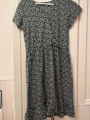 Svart blommig klänning XOXO Friends - Söt svart klänning från XOXO Friends med vitt blommönster. Klänningen har korta ärmar, v-ringning och en avslappnad passform. Perfekt för sommarens alla tillfällen och enkel att styla med sneakers eller sandaler.