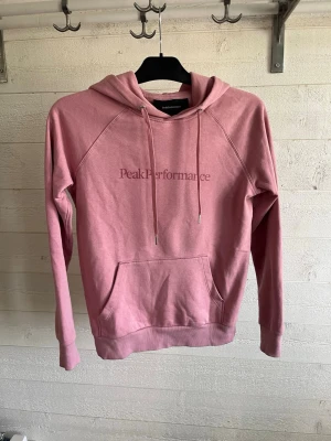 Rosa hoodie från Peak Performance - Säljer en snygg rosa hoodie från Peak Performance med huva och dragsnören. Hoodien har en stor magficka och logga broderad på bröstet. Tillverkad i mjukt bomullsmaterial och har ribbade muddar vid ärmar och nederkant.