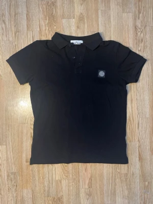 Svart pikétröja med krage - Klassisk svart pikétröja med korta ärmar och krage. Tröjan har knappar framtill och en diskret logga på bröstet. Perfekt till jeans eller shorts för en clean och enkel look. Skick: Väldigt fint skick och inga defekter. Inga hål eller andra tydliga skador. 