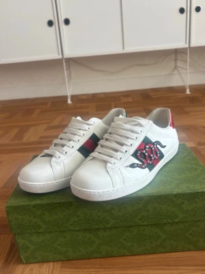 Gucci vita sneakers med orm - Snygga vita sneakers från Gucci i läder med den ikoniska gröna och röda sidoremsan samt en broderad orm på sidan. Baksidan har röd och grön metallicdetalj och skorna har vita snören. Perfekta för dig som vill sticka ut med lyxig streetstyle. Skorna är använda 6 gånger pris kan diskuteras fråga om det är något ni undrar.