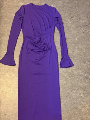 Lång lila klänning med trumpetärm - Snygg långklänning i stark lila färg med långa ärmar och trumpetformade ärmslut. Klänningen har en åtsittande passform och draperad detalj i midjan som ger en cool twist. Perfekt för dig som vill sticka ut med en unik look.