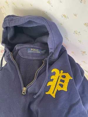 Blå hoodie från Polo Ralph Lauren - Mörkblå hoodie från Polo Ralph Lauren med halv dragkedja och stor gul broderad bokstav på bröstet. Klassisk känguruficka framtill och justerbar huva med snören. Tillverkad i mjuk bomull för skön känsla. Om ni har funderingar så svarar jag gärna.