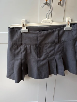 Mörkgrå plisserad minikjol - Snygg mörkgrå kortkjol med plisserade veck och bred linning. Kjolen har hällor för bälte och är tillverkad i ett mjukt bomullsliknande material. Perfekt för en trendig och avslappnad stil.