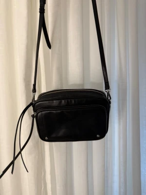 Snygg handväska - Snygg svart axelväska i skinnimitation med justerbart axelband och två dragkedjefack. Väskan har en stilren design med silverfärgade detaljer och dekorativa remmar på sidan. Perfekt storlek för det viktigaste och passar till många olika outfits.