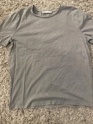Grå basic t-shirt från Zara - En enkel grå t-shirt från Zara med rund halsringning och korta ärmar. Tillverkad i mjukt bomullstyg som känns skönt mot huden. Perfekt till jeans eller shorts för en clean och avslappnad stil.