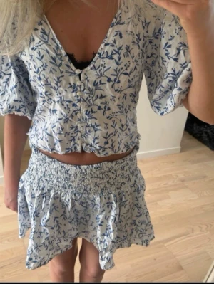 Vit blommig blus med puffärm - Söt kort blus i vitt med blått blommönster och puffiga trekvartsärmar. Blusen har v-ringning, knappar framtill och en lätt croppad passform. Perfekt till sommaren och ger en fräsch, romantisk look.