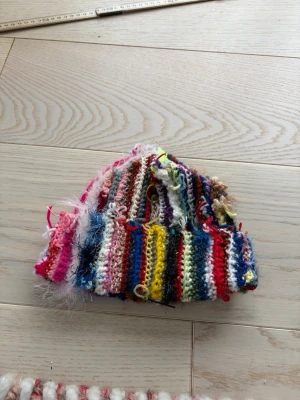 Flerfärgad handvirkad  mössa - Unik och färgglad handvirkad mössa med ränder i rosa, blått, gult, rött, brunt och vitt. Mössan har en kreativ och bohemisk look med olika garnsorter och texturer, inklusive fluffiga detaljer. Perfekt för dig som vill sticka ut och gillar personliga accessoarer.Höjd ca 25 cm  Omkrets ca 48 cm