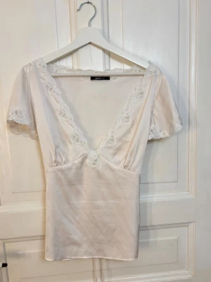 Gina Tricot Superpopulär Satin Lace Top – vit - Superpopulär Satin Lace Top från Gina Tricot i vit satin med fina spetsdetaljer och knytning baktill. Märkt strl 44, men skulle säga att den passar mer som L då den är justerbar. Obs! Den ursprungliga knytningen framtill är borttagen på denna topp. Det finns även en suuuupersvag fläck på framsidan, som syns på bild 4. Den syns knappt när toppen bärs och fanns redan när jag köpte toppen på Vinted. Kan eventuellt gå bort vid ordentlig tvätt. 