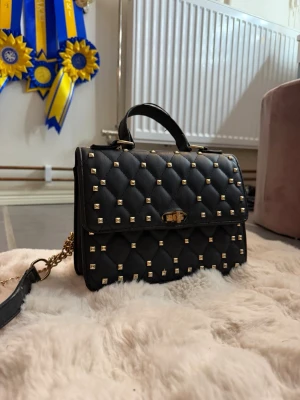 Svart quiltad handväska med nitar - Snygg svart handväska i quiltad design med guldfärgade nitar och kedjedetaljer. Väskan har ett topphandtag, vridlås framtill och flera fack inuti, inklusive ett med dragkedja. Perfekt accessoar för att lyfta din outfit. Lite sliten och välanvänd men ok skick.