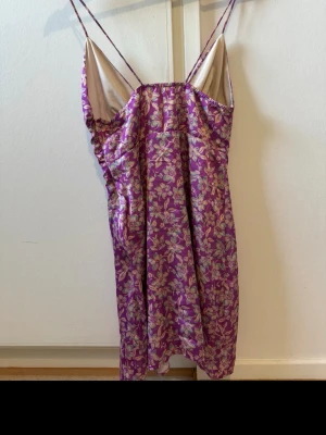 Lila blommigt klänning med snörning - Sött klänning i lila med blommigt mönster och tunna axelband. Klänningen har snörning framtill och är gjort i ett lite glansigt, mjukt material som känns lyxigt mot huden. Perfekt för mysiga kvällar 