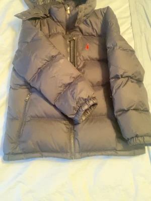 Ralph lauren Svart dunjacka med huva - Snygg svart dunjacka med huva och dragkedja framtill. Jackan har två sidofickor med dragkedja och en bröstficka. Puffig modell med elastiska muddar vid ärmslut. Röd logga broderad på bröstet.