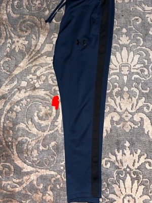 under armour byxor  - Snygga marinblå träningsbyxor från Under Armour med svarta ränder längs sidorna. Byxorna har dragsko i midjan och smal passform. Perfekta för dig som gillar sportig stil och vill ha bekväma byxor till vardagen.