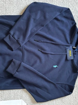 Mörkblå hoodie från Polo Ralph Lauren - Snygg mörkblå hoodie från Polo Ralph Lauren med klassisk huva och dragsko. Har den ikoniska broderade loggan i ljusblått på bröstet och en stor magficka. Tillverkad i mjukt bomullsmaterial och har ribbade muddar vid ärmar och nederkant.