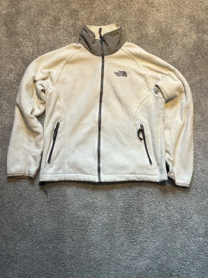 Beige fleecejacka från The North Face - Mysig beige fleecejacka från The North Face med hög krage och hel dragkedja framtill. Jackan har två fickor med dragkedja och detaljer i grått vid kragen. Perfekt för kyliga dagar och enkel att matcha med olika outfits.