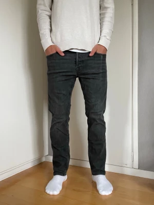 Jack & Jones Jeans - Snygga svart/grå jeans från Jack & Jones i storleken 29/30. Modellen på jeansen heter TAPERED/MIKE och är straight men smalnar av något mot slutet. Jag på bilden är ca 178 cm och väger ca 70 kg och jeansen är något för små för mig. På sista bilden kan man se en liten slitning på vänster ficka där jag har haft mobilen. Tveka inte på att höra av dig vid minsta fundering!